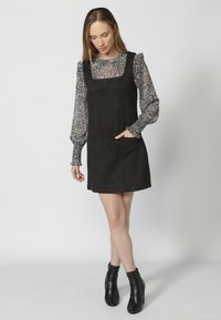 Robe en daim noir avec un décolleté carré et des poches avant, superposée à un blouse à manches longues à motifs ; associée à des bottines noires.