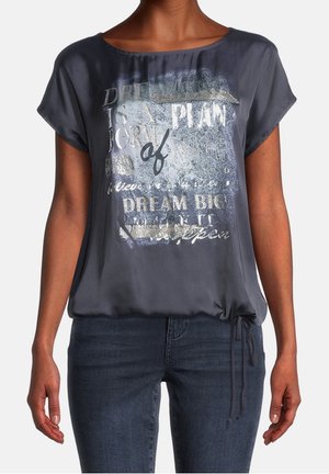 Vrouw die een donkergrijze korte mouwen blouse met metalen tekst en donkerblauwe spijkerbroek draagt, staand met armen ontspannen langs haar zij.