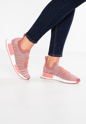 Person iført lyserøde strikkede sneakers med hvide såler og koralfarvede detaljer, kombineret med mørkeblå skinny jeans på hvid baggrund.