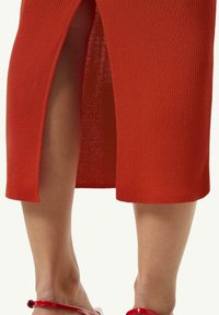 Gestreiftes rotes Midikleid mit seitlichem Schlitz, das eine strukturierte Passform und eine glatte Textur aufweist. Getragen mit glänzenden roten Sandalen.