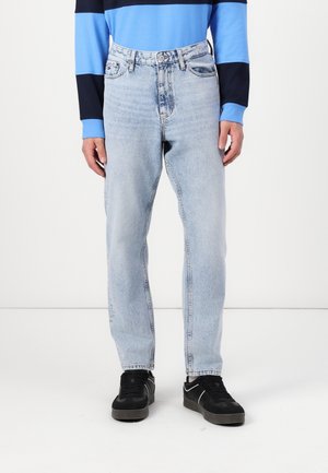 Person trägt hellblaue Jeans, ein langärmeliges Hemd mit blauen und schwarzen Streifen und schwarze Sneaker mit weißen Akzenten.