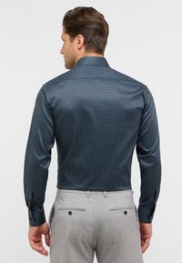 Camicia a maniche lunghe di colore blu scuro con piccoli pois bianchi, texture liscia e polsini con bottoni, abbinata a pantaloni grigi chiari.