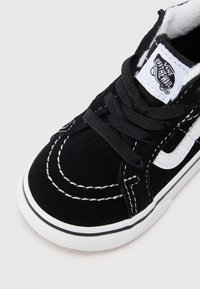 Crne Vans tenisice s bijelim šavovima, vezicama i gumenim potplatom, s Vans Off The Wall logom na jeziku.