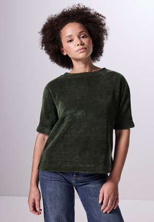 Top in velluto a coste verde scuro con maniche corte e scollatura ampia, abbinato a jeans in denim azzurro chiaro. Il tessuto presenta una finitura strutturata.