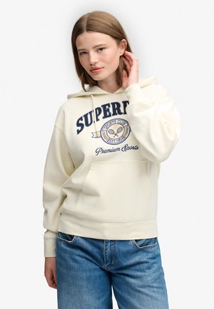 Ung kvinde med glat lyst brunt hår iført en cremefarvet hættetrøje med sportslogo og blå jeans, poserer med hånden tæt på øret.