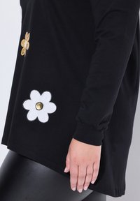 Maglietta nera a maniche lunghe con applicazioni floreali bianche e dorate sul davanti inferiore, orlo arrotondato e polsini a costine.
