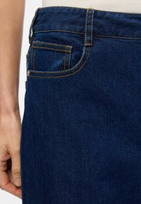 Σκούρες μπλε τζιν παντελόνες με κίτρινη ραφή, με πίσω τσέπη και λεία υφή. Κοντινό πλάνο της περιοχής της μέσης.