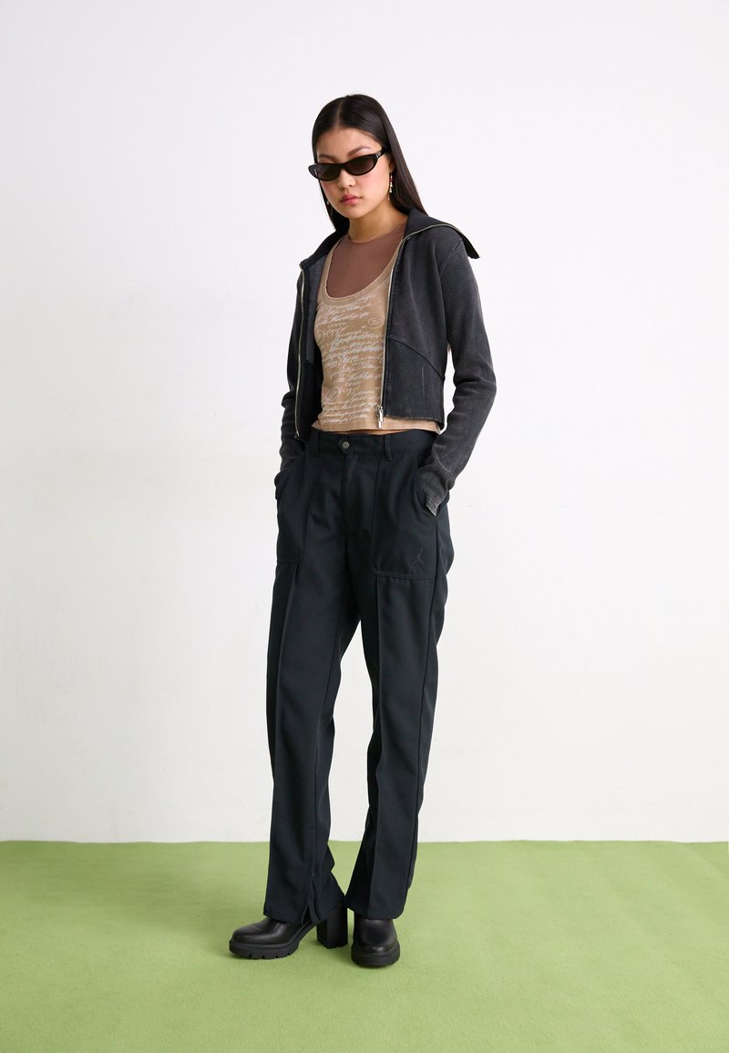 Schwarze, cropped Zip-Up-Trainingsjacke, beiges Tanktop mit Text, dunkle Hose mit Taschen und klobigen schwarzen Plateaustiefeln. Glattes Material, entspannte Passform.