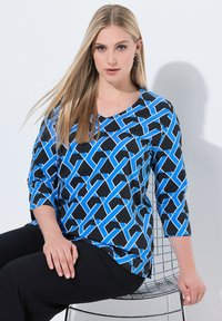 Maglietta a maniche 3/4 nera con un motivo geometrico blu. Tessuto morbido con scollatura a V. Il modello è seduto su una sedia in metallo.