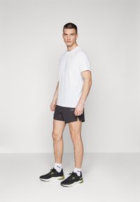 Camiseta blanca de manga corta, pantalones cortos negros, zapatillas negras y verdes. Diseño minimalista con logotipos sutiles en la camiseta y los pantalones cortos.