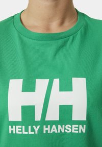 Zelené bavlněné tričko s kulatým výstřihem, se symbolem velkého bílého loga "HH" a textem "HELLY HANSEN" pod ním ve výrazném bílém písmu.