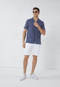 Chemise à rayures à manches courtes en tissu bleu marine et blanc, boutonnée, associée à un short blanc et des baskets blanches, se tenant devant un mur en briques claires.
