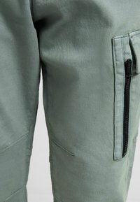 Gröna cargobyxor med en slät textur, sidodragkedjeficka och vinklade sömmar på benen.