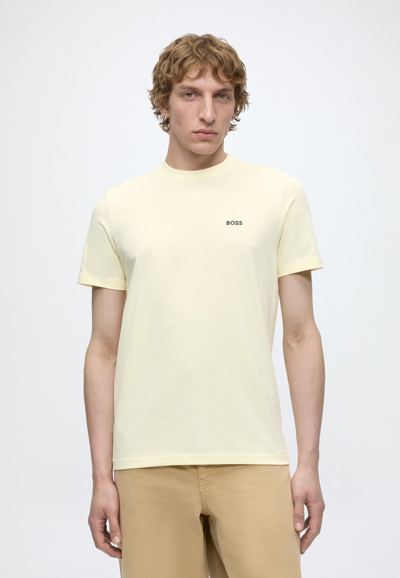 Jeune homme aux cheveux blonds bouclés portant un t-shirt BOSS jaune clair et un pantalon beige, debout devant un fond blanc uni.
