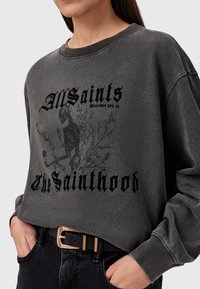 Donna che indossa una felpa nera sbiadita con il testo gotico "AllSaints ti dà il benvenuto nel Sainthood" e una grafica di uno scheletro con rami.