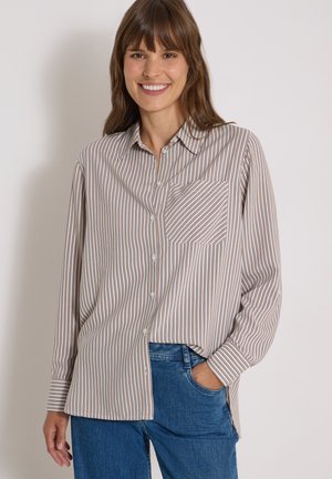 Kvinde smilende iført en langærmet beige og brunstribet skjorte med knapper og en brystlomme samt blå jeans, hånd i lommen.