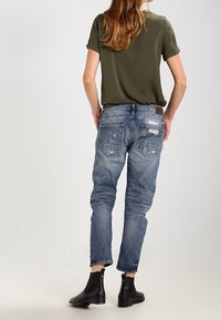 Femme avec de longs cheveux portant un t-shirt vert olive, un jean bleu déchiré et des bottines noires, se tenant de dos sur fond uni.