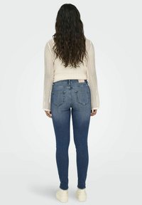 Jeans ajustados en denim azul oscuro con una cintura alta, que cuentan con cinco bolsillos y detalles de costura. Combinados con un suéter texturizado en crema.