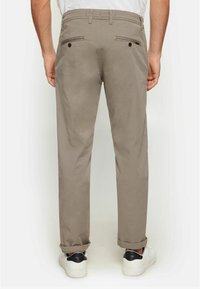 Beige Chino-Hose mit geradem Schnitt, zwei Gesäßtaschen, die mit Knöpfen gesichert sind, und umgeschlagenen Säumen. Getragen mit weißen Sneakers.