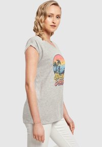 Grå t-skjorte med korte ermer som har et fargerikt grafisk design med palmer og teksten "Stranger Things." Glatt bomullsstoff, avslappet passform.
