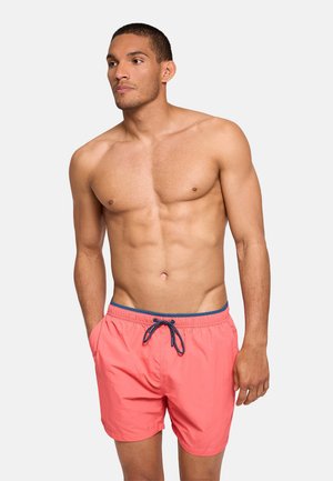 Hombre sin camisa con pelo corto que lleva pantalones cortos de baño color coral con cordón azul marino, de pie con las manos en los bolsillos frente a un fondo liso.
