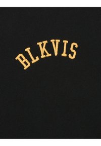 Mustat T-paita, jossa on kaareva keltainen brodeerattu teksti "BLKVIS" keskitettynä yläpuolelle. Tasainen kangas, rento muotoilu.
