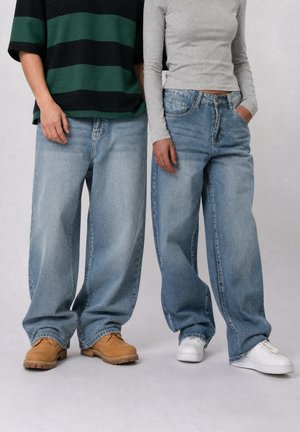 Deux personnes portant des jeans bleus larges et des hauts décontractés, l'une en bottes marron et l'autre en baskets blanches, debout côte à côte.