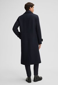 Marineblauer Wollmantel mit geradem Schnitt, großem Kragen und seitlichen Taschen. Hat einen einzelnen Rückenschlitz und knöpfbare Manschetten. Schwarze Stiefel sichtbar.