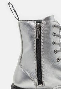 Botte en cuir argenté avec fermeture éclair latérale noire étiquetée "Dr. Martens", lacets gris, et une boucle en tissu à l'arrière.