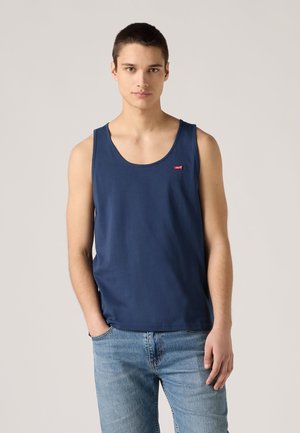 Mladý muž v tmavomodrom tielku bez rukávov a svetlomodrých džínsoch, stojaci s jednou rukou v vrecku na jednoduchom pozadí.