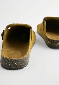 Brune ruskinds slip-on sko med en tekstureret sort plettet sål. Designet har en rund form og en justerbar rem på siden.