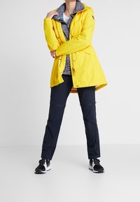 Osoba w żółtej kurtce przeciwdeszczowej, szarej bluzie z zamkiem, czarnych spodniach i czarno-białych trampkach stoi na białym tle.