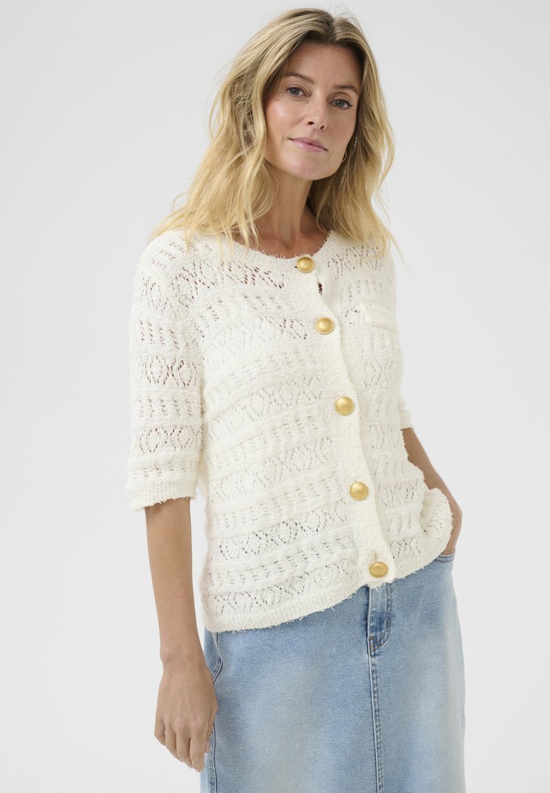 Witte gebreide cardigan met een textuurontwerp, voorzien van korte mouwen, ronde halslijn en gouden knopen. Draagt over een lichtblauwe denimrok.