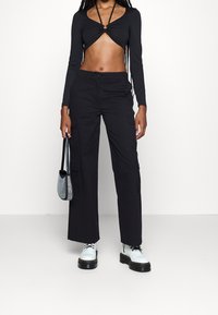 Crop top noir à manches longues avec un devant torsadé, associé à un pantalon cargo noir à jambes larges avec poches latérales. Le mannequin tient un petit sac argenté.