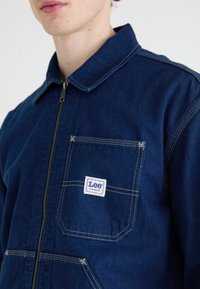 Joven vistiendo una chaqueta de mezclilla azul oscuro con cremallera y un bolsillo en el pecho etiquetado "Lee Workwear" con costuras en contraste.