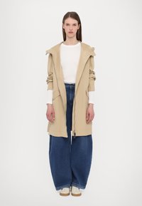 Giacca lunga beige con cappuccio, dotata di zip frontale e tasche, indossata sopra una maglia bianca e pantaloni in denim blu a gamba larga con vestibilità rilassata.