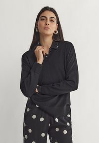 Pull en tricot noir avec un col en V, accompagné d'une chemise à col à pois contrastants en dessous. Texture douce et coupe décontractée.