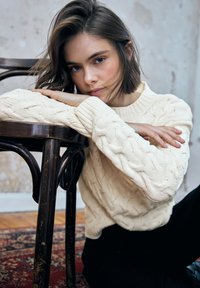 Kremowy sweter z dzianiny warkoczowej z prążkowanym dekoltem, noszony z ciemnymi spodniami. Modelka opiera się o drewniane krzesło w teksturowanym wnętrzu.