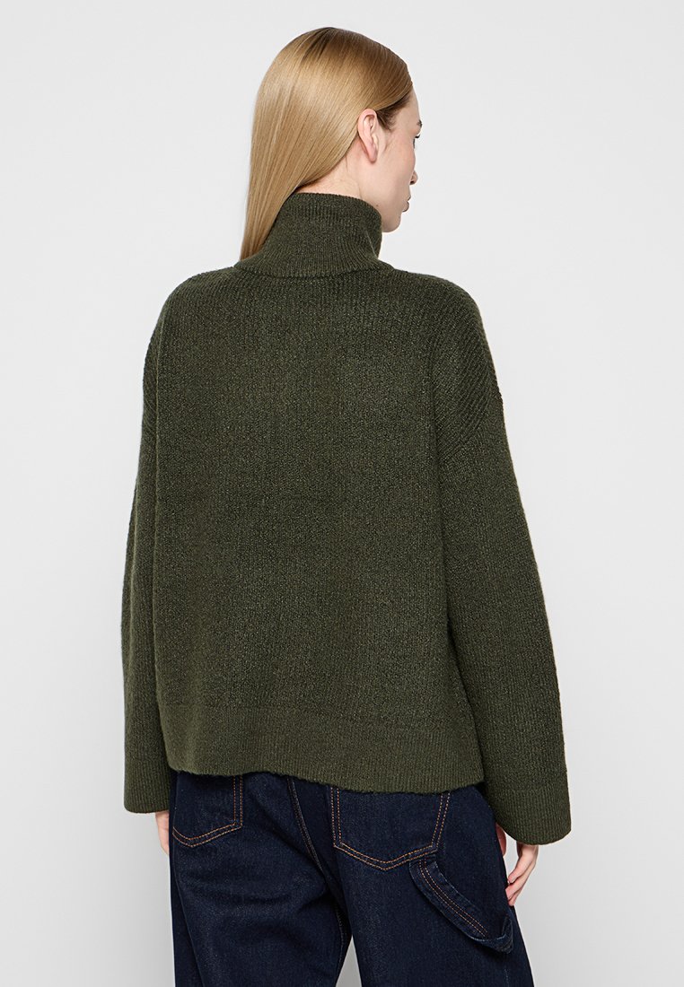 Dunkelgrüner, gestrickter Pullover mit hohem Kragen, lockerer Passform und gerippten Details, kombiniert mit blauen Jeans. Rückansicht dargestellt.