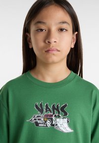 Grünes Baumwoll-T-Shirt mit einem grafischen Design eines Oldtimers und einer Skateboardrampe, versehen mit dem Text "VANS" in stilisierten schwarzen Buchstaben.