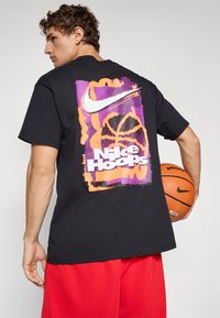 Jeune homme tenant un ballon de basket, portant un t-shirt noir avec un graphique coloré "Nike Hoops" et un short rouge, se tenant devant un fond uni.