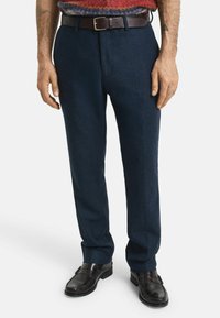 Marineblaue Tailor-Trousers aus strukturiertem Stoff mit geradem Schnitt, kombiniert mit einem braunen Ledergürtel. Schwarze Slipper runden den Look ab.
