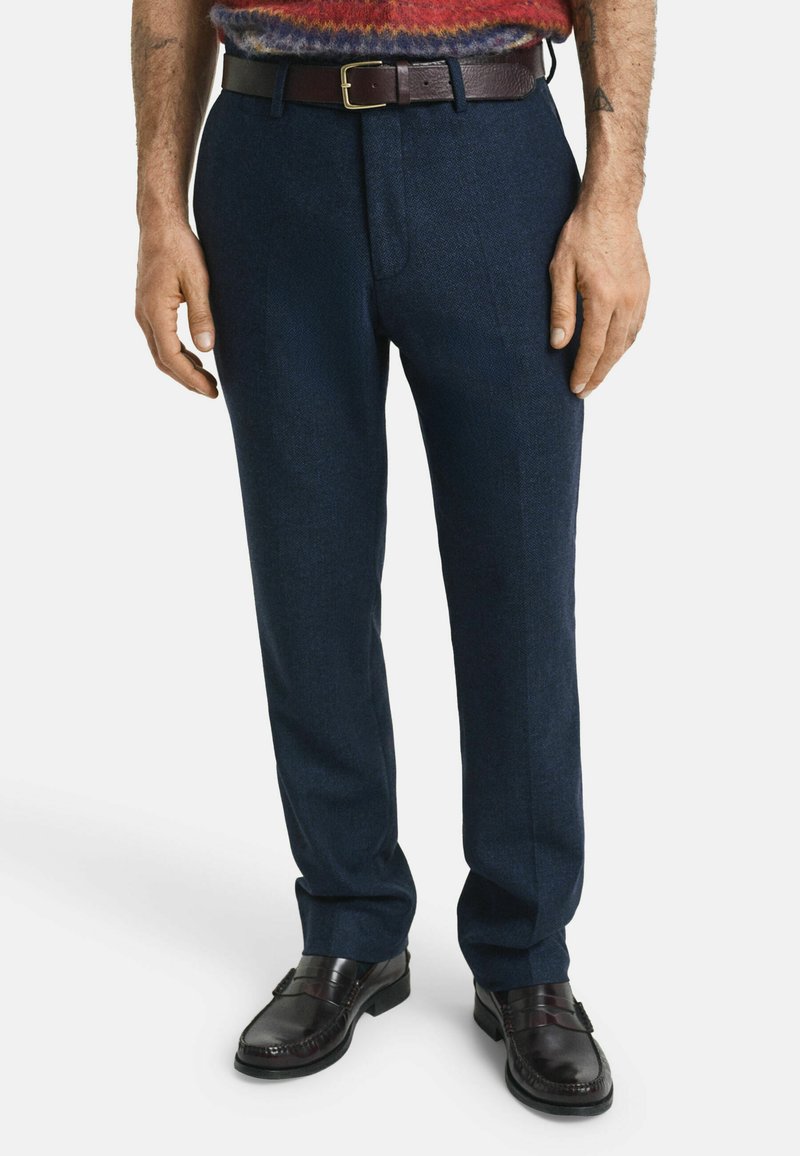 Marineblaue Tailor-Trousers aus strukturiertem Stoff mit geradem Schnitt, kombiniert mit einem braunen Ledergürtel. Schwarze Slipper runden den Look ab.