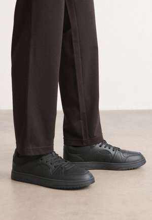 Personne portant des baskets noires à lacets et un pantalon droit marron foncé debout sur un sol clair.