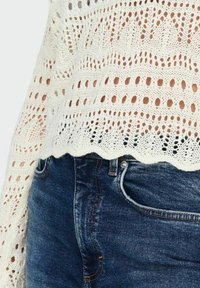 Top bianco lavorato a uncinetto a maniche lunghe con orlo smerlato abbinato a jeans di denim blu, focalizzato sull'area della vita e del ventre.