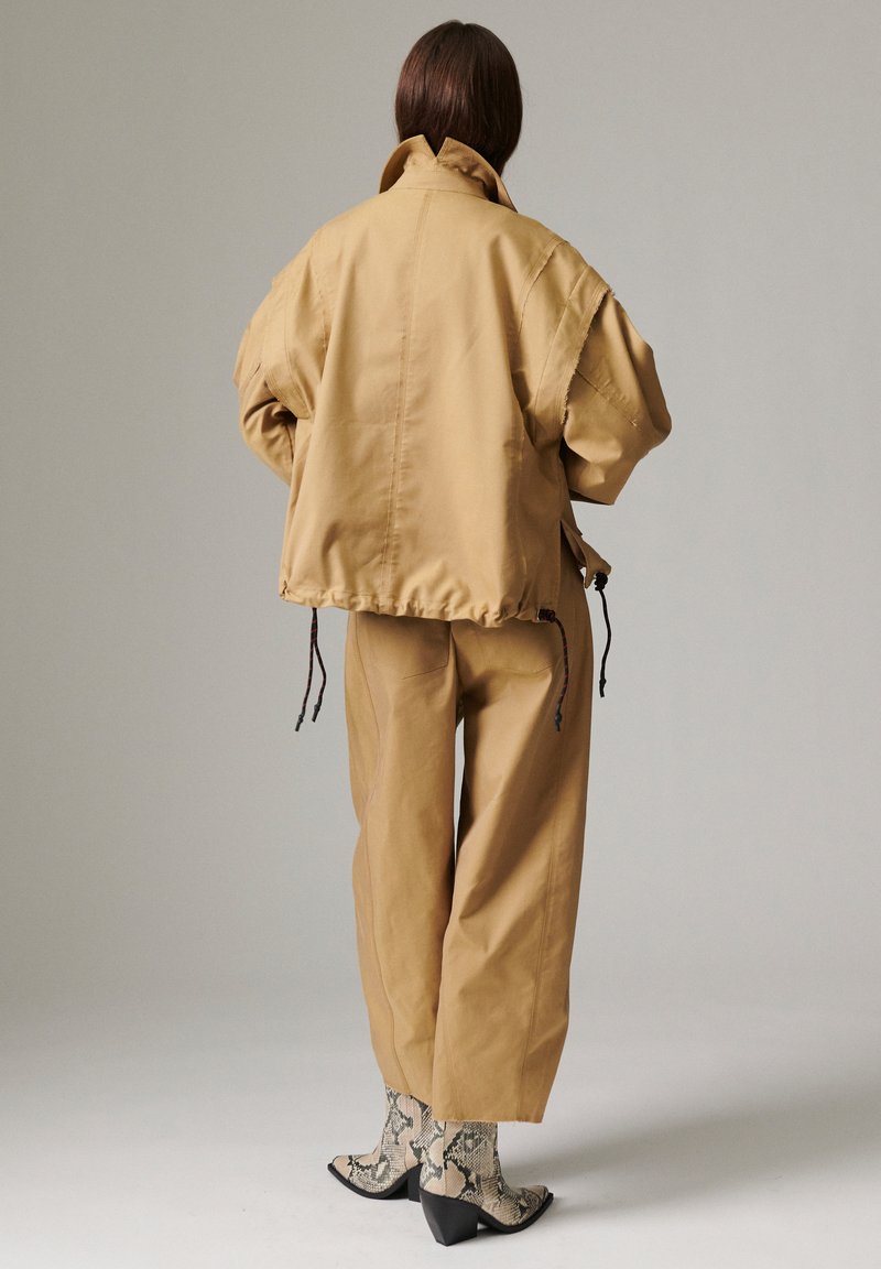 Άτομο με καστανά μαλλιά που φοράει χαλαρό μπεζ μπουφάν και παντελόνια, συνδυασμένα με ασπρόμαυρα μποτάκια με σχέδιο φίδι, στέκεται πλάτη.