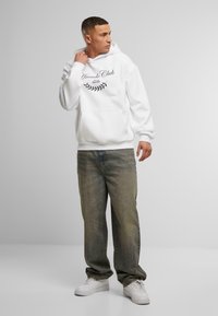 Felpa bianca con cappuccio, dotata di tasca frontale e design testuale nero. Indossata con jeans verdi sbiaditi e sneakers bianche. Tessuto dalla texture liscia.