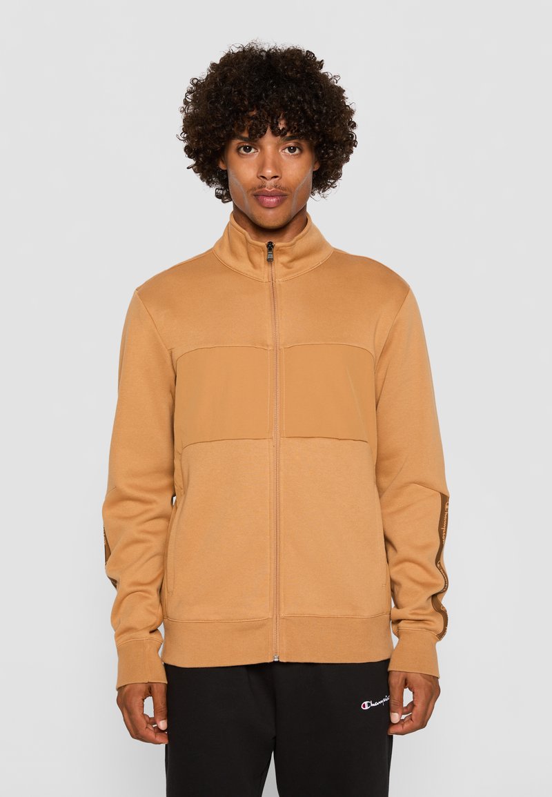Tan zip-up jakke med høj krave, der har et solidt brystpanel og accentstriber på ærmerne. Lavet af et blødt stof, parret med sorte bukser.