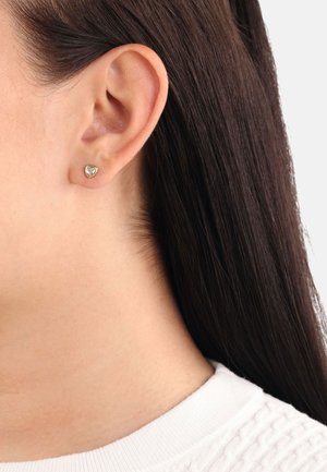 Pendiente de aro en forma de corazón en tono dorado con una gema transparente en el centro. Colocado sobre cabello liso y recto de color oscuro y un suéter claro.