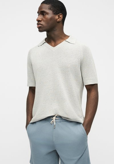 Homme portant un haut en maille à manches courtes beige clair avec un col et un short bleu à cordon de serrage, les mains dans les poches, regardant sur le côté.